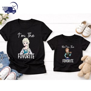 Puede incluir: Dos camisetas negras con texto blanco e im&aacute;genes de dibujos animados. Una camiseta dice "I'm The FUVORITE" con una imagen de Elsa. La otra dice "No, I'm The FUVORITE" con Anna. Zapatos blancos y un peluche tambi&eacute;n est&aacute;n en la imagen.