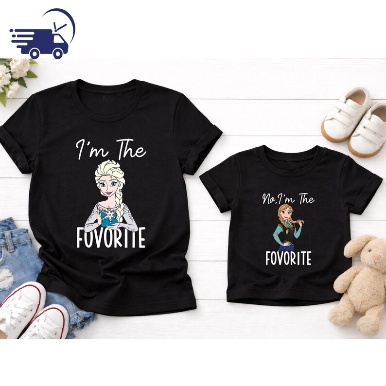 Puede incluir: Dos camisetas negras con texto blanco e im&aacute;genes de dibujos animados. Una camiseta dice "I'm The FUVORITE" con una imagen de Elsa. La otra dice "No, I'm The FUVORITE" con Anna. Zapatos blancos y un peluche tambi&eacute;n est&aacute;n en la imagen.