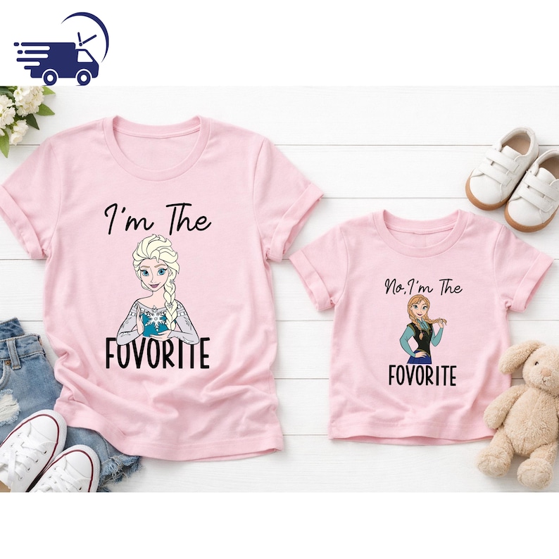 Puede incluir: Dos camisetas rosas con im&aacute;genes de dibujos animados de Elsa y Anna de Frozen. La camiseta m&aacute;s grande dice "I'm The FAVORITE" y la m&aacute;s peque&ntilde;a dice "No, I'm The FAVORITE". Zapatillas blancas, un peluche y pantalones cortos de mezclilla en el fondo.