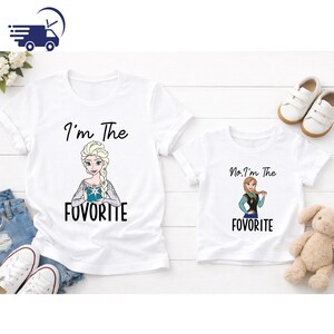 Puede incluir: Dos camisetas blancas con im&aacute;genes de dibujos animados de Elsa y Anna de Frozen. La camiseta de adulto dice "I'm The FovoritE" y la camiseta infantil dice "No, I'm The FovoritE". Las camisetas est&aacute;n sobre una superficie de madera blanca.