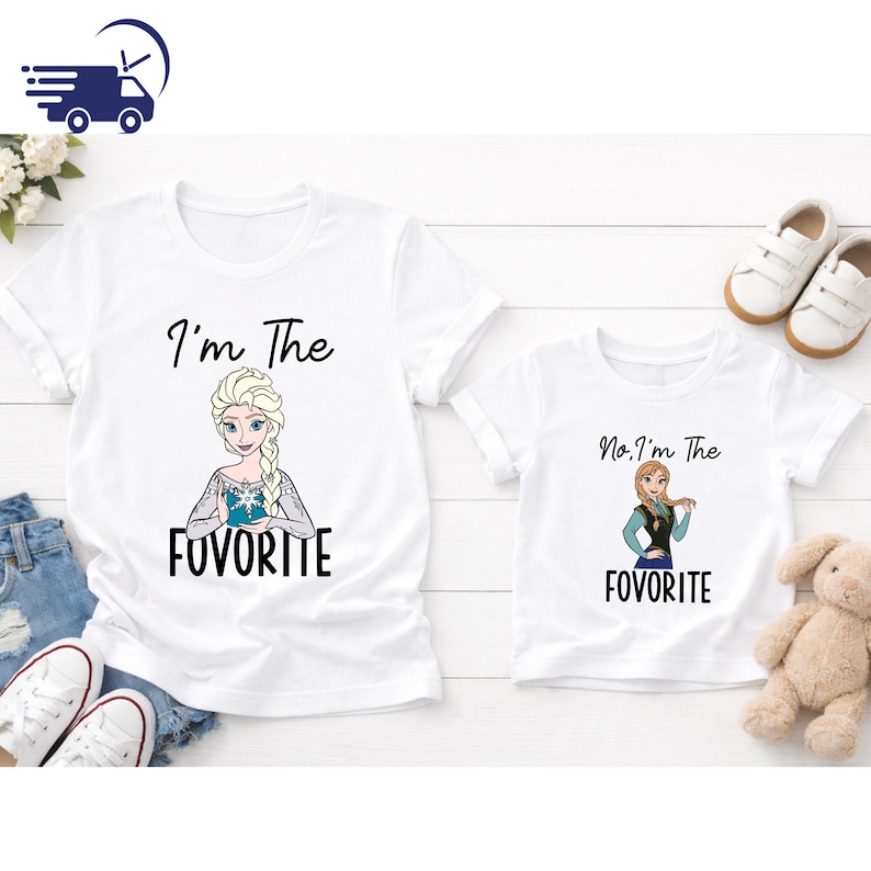Puede incluir: Dos camisetas blancas con im&aacute;genes de dibujos animados de Elsa y Anna de Frozen. La camiseta de adulto dice "I'm The FovoritE" y la camiseta infantil dice "No, I'm The FovoritE". Las camisetas est&aacute;n sobre una superficie de madera blanca.