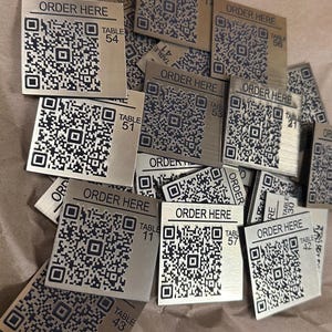May include: A collection of square, metallic-finish signs featuring a black QR code, the text "ORDER HERE" at the top, and a specific table number printed on the right side. Each sign is distinct, displaying various numbers such as 54, 51, 11, 57, 43, 56, 21, 30, and 42.