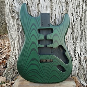 Puede incluir: Un cuerpo de guitarra verde con un patr&oacute;n de vetas de madera &uacute;nico. El cuerpo tiene recortes para pastillas, controles y un sistema de tr&eacute;molo. La veta de la madera es verde oscuro con acentos negros. El cuerpo de la guitarra est&aacute; sin terminar.