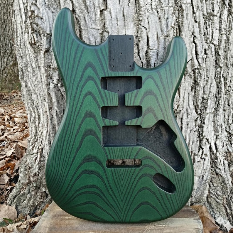 Puede incluir: Un cuerpo de guitarra verde con un patr&oacute;n de vetas de madera &uacute;nico. El cuerpo tiene recortes para pastillas, controles y un sistema de tr&eacute;molo. La veta de la madera es verde oscuro con acentos negros. El cuerpo de la guitarra est&aacute; sin terminar.