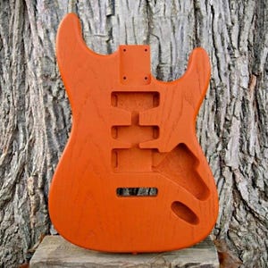 May include: An orange guitar body with cutouts for pickups, a tremolo system, and neck attachment. The unfinished wood has a visible grain and is set against a textured tree trunk background. The body is designed for an electric guitar.