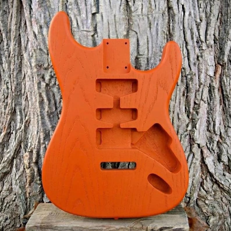 May include: An orange guitar body with cutouts for pickups, a tremolo system, and neck attachment. The unfinished wood has a visible grain and is set against a textured tree trunk background. The body is designed for an electric guitar.