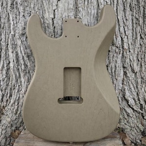 May include: A beige guitar body made of wood, featuring cutouts for pickups, a tremolo system, and neck attachment. The unfinished body has a smooth, curved design, ready for customization. The guitar body is shown against a textured tree bark background.
