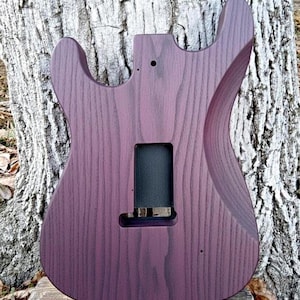 Puede incluir: Cuerpo de guitarra de madera te&ntilde;ida de p&uacute;rpura con forma contorneada. El cuerpo presenta cortes para acceder al m&aacute;stil y una cavidad rectangular para la electr&oacute;nica. La veta de la madera es visible y el acabado es liso.