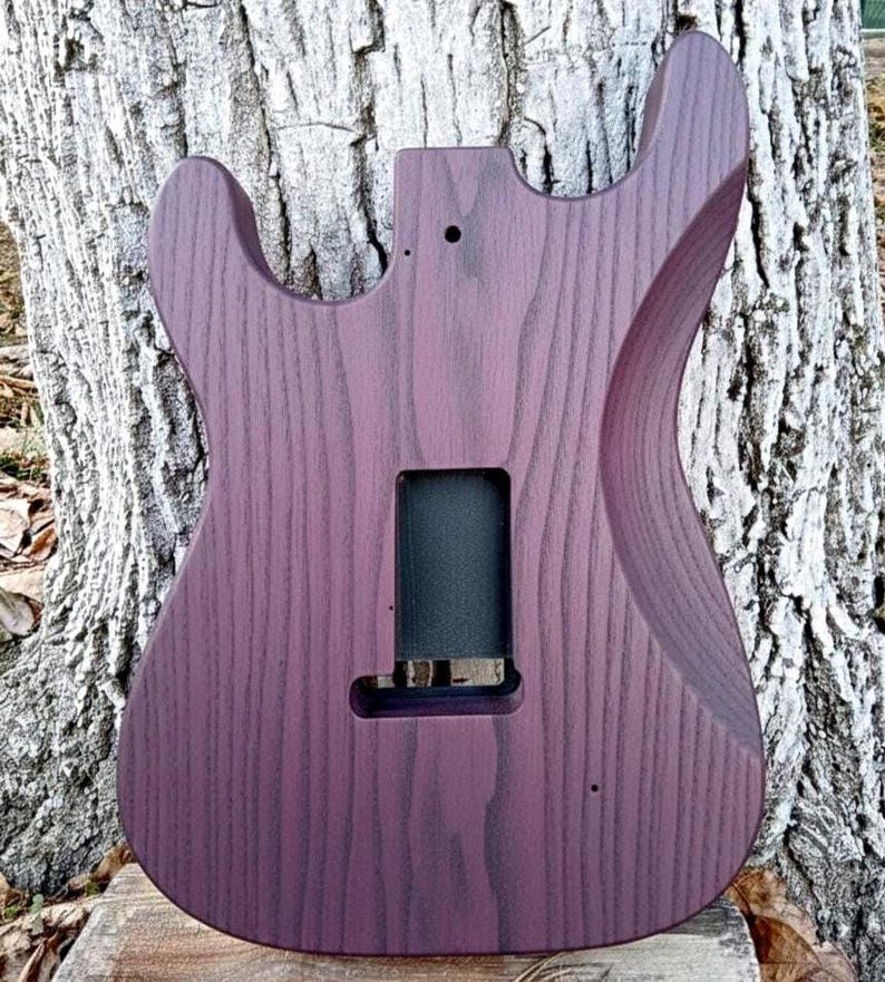 Puede incluir: Cuerpo de guitarra de madera te&ntilde;ida de p&uacute;rpura con forma contorneada. El cuerpo presenta cortes para acceder al m&aacute;stil y una cavidad rectangular para la electr&oacute;nica. La veta de la madera es visible y el acabado es liso.