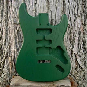 May include: A solid green guitar body with cutouts for pickups, controls, and a tremolo system. The unfinished wood has a visible grain and is set against a textured tree trunk background. The body is designed for a bolt-on neck.