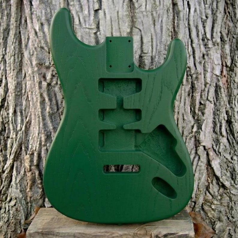 May include: A solid green guitar body with cutouts for pickups, controls, and a tremolo system. The unfinished wood has a visible grain and is set against a textured tree trunk background. The body is designed for a bolt-on neck.