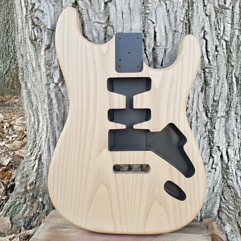 May include: Unfinished electric guitar body made of light-colored wood. The body has cutouts for pickups, controls, and a neck. A black metal plate is attached at the top for the neck. The wood grain is visible, giving it a natural look.