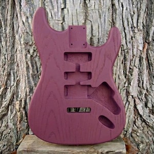 May include: A solid body electric guitar body in a deep burgundy color. The wood grain is visible. The body has cutouts for pickups, controls, and a tremolo system. The guitar body is unfinished.