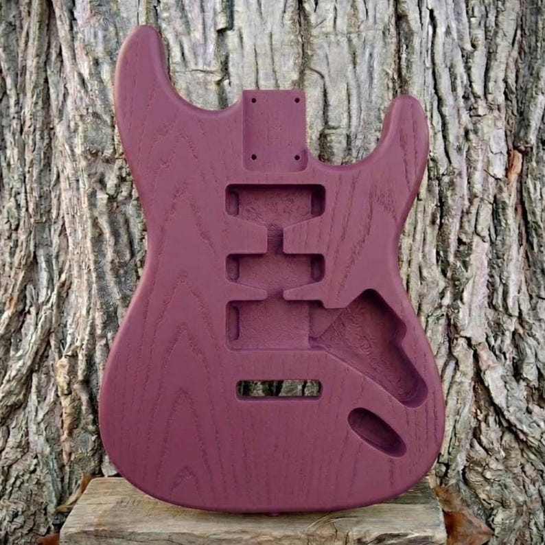 May include: A solid body electric guitar body in a deep burgundy color. The wood grain is visible. The body has cutouts for pickups, controls, and a tremolo system. The guitar body is unfinished.