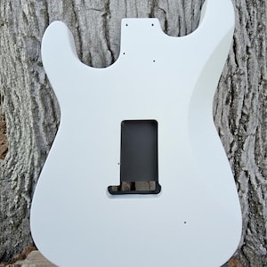 Puede incluir: Un cuerpo de guitarra el&eacute;ctrica blanco con huecos para pastillas y controles. El cuerpo es liso y pintado de blanco, con una cavidad rectangular para el puente y otra m&aacute;s peque&ntilde;a para la pastilla del m&aacute;stil. El cuerpo de la guitarra est&aacute; sobre un fondo de madera texturizada.