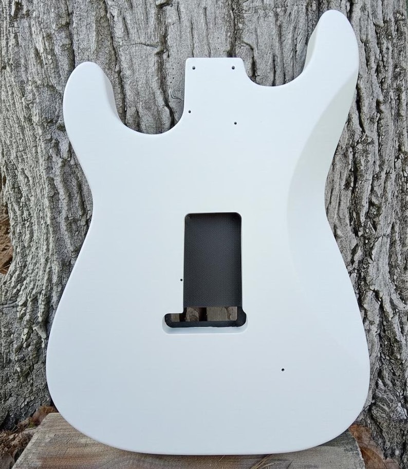 Puede incluir: Un cuerpo de guitarra el&eacute;ctrica blanco con huecos para pastillas y controles. El cuerpo es liso y pintado de blanco, con una cavidad rectangular para el puente y otra m&aacute;s peque&ntilde;a para la pastilla del m&aacute;stil. El cuerpo de la guitarra est&aacute; sobre un fondo de madera texturizada.