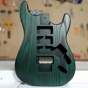 Könnte beinhalten: Ein grüner und schwarzer E-Gitarrenkorpus mit Holzmaserung. Der Korpus hat Aussparungen für Tonabnehmer, ein Tremolo-System und einen Hals. Die Oberfläche ist dunkelgrün mit schwarzen Akzenten. Der Gitarrenkorpus ist unfertig.