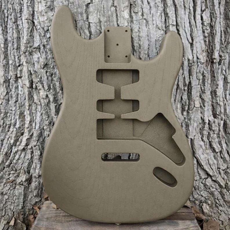 May include: A tan-colored, unfinished electric guitar body. The body has cutouts for pickups, controls, and a neck. The wood grain is visible. The guitar body is designed to be customized.