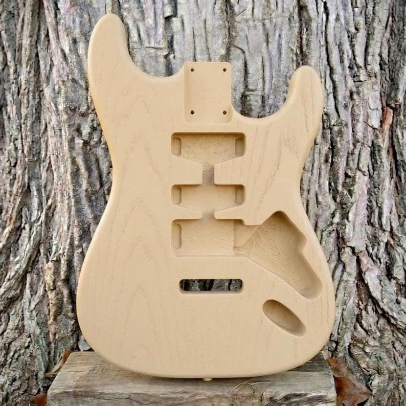 May include: Unfinished electric guitar body made of light-colored wood. The body has cutouts for pickups, controls, and a tremolo system. The wood grain is visible, and the body is ready for finishing.