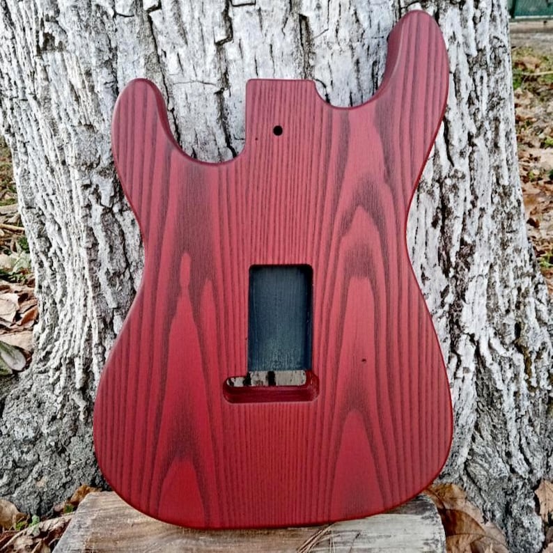 Puede incluir: Un cuerpo de guitarra rojo con un acabado de veta de madera natural. El cuerpo tiene recortes para el m&aacute;stil, las pastillas y otros herrajes. La veta de la madera es visible y el acabado es liso.