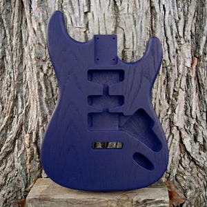 May include: A solid navy blue electric guitar body, unfinished, with cutouts for pickups, controls, and neck. The wood grain is visible. The guitar body is set against a textured tree trunk background.
