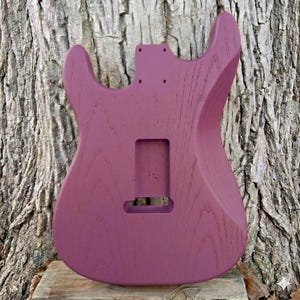 May include: A solid purple guitar body with a smooth finish. The body has cutouts for the neck, pickups, and other hardware. The wood grain is visible. The guitar body is set against a textured tree trunk background.