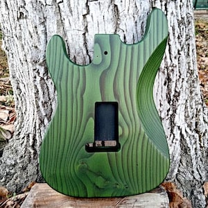 Puede incluir: Un cuerpo de guitarra de madera verde y marr&oacute;n con un patr&oacute;n de vetas &uacute;nico. El cuerpo tiene recortes para el m&aacute;stil, las pastillas y el puente. El cuerpo de la guitarra est&aacute; sin terminar.