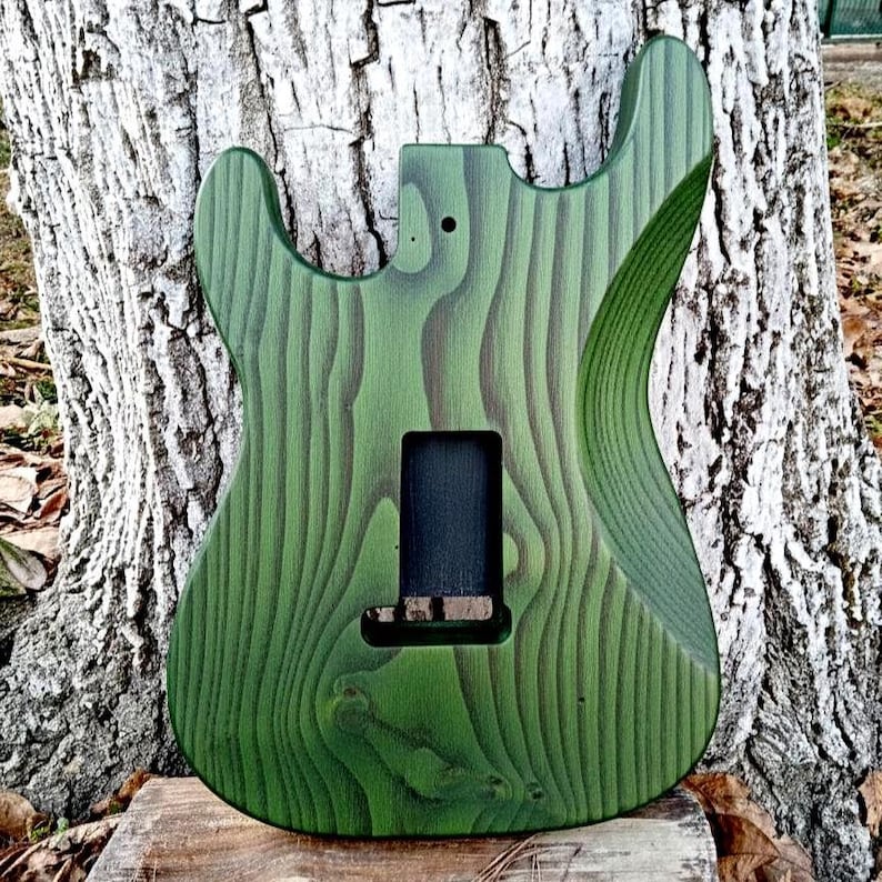 Puede incluir: Un cuerpo de guitarra de madera verde y marr&oacute;n con un patr&oacute;n de vetas &uacute;nico. El cuerpo tiene recortes para el m&aacute;stil, las pastillas y el puente. El cuerpo de la guitarra est&aacute; sin terminar.