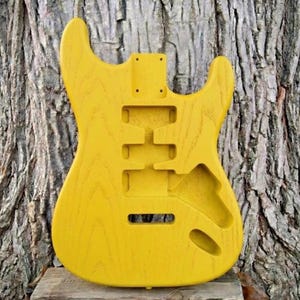 May include: A bright yellow, unfinished electric guitar body. The body has cutouts for pickups, controls, and a neck. The wood grain is visible, and the body is set against a textured tree trunk background. This is a DIY guitar project.