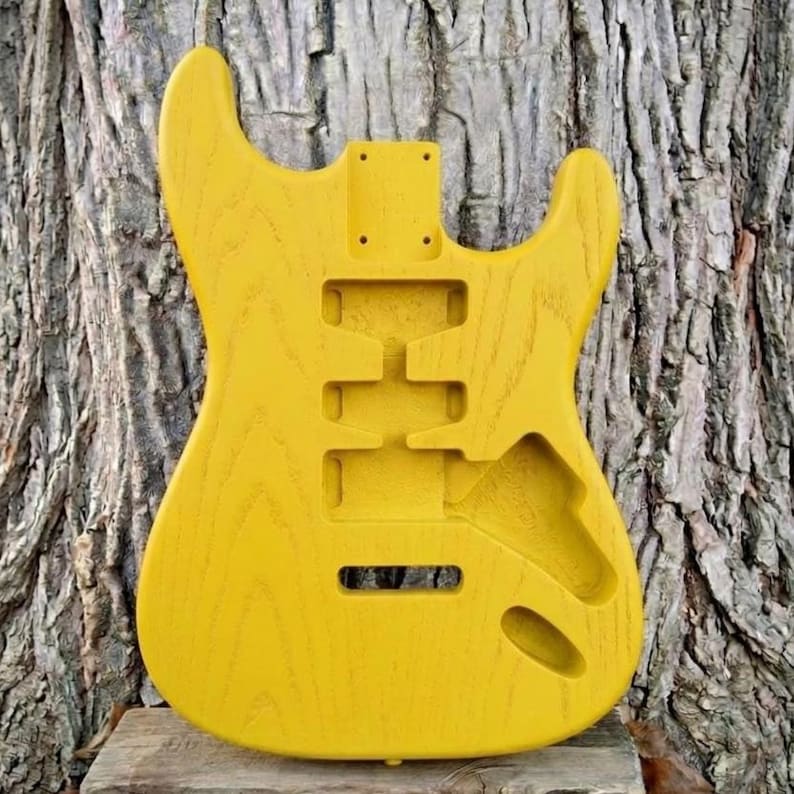 May include: A bright yellow, unfinished electric guitar body. The body has cutouts for pickups, controls, and a neck. The wood grain is visible, and the body is set against a textured tree trunk background. This is a DIY guitar project.