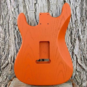 May include: An orange electric guitar body with a wood grain finish. The body has cutouts for the neck, pickups, and other hardware. The guitar body is set against a textured tree trunk background.
