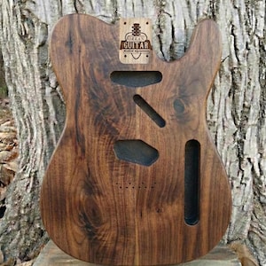 Może przedstawiać: Korpus gitary z ciemnobrązowego drewna, z wycięciami na przetworniki i elementy sterujące. Główka posiada logo "ORKAN GUITAR". Widoczne usłojenie drewna, korpus jest niewykończony.