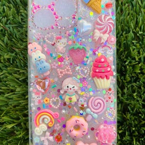 May include: Clear phone case decorated with various colorful, 3D embellishments. The case features a variety of charms, including a cupcake, ice cream cone, bow, donut, and a rainbow. The word "ROSY" is visible on the case.