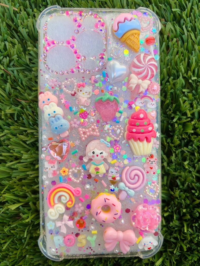 May include: Clear phone case decorated with various colorful, 3D embellishments. The case features a variety of charms, including a cupcake, ice cream cone, bow, donut, and a rainbow. The word "ROSY" is visible on the case.
