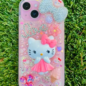 May include: Pink phone case with a Hello Kitty design. The case features a large, sparkling bow, the word "Love" in rhinestones, and various decorative elements like flowers, hearts, and an ice cream cone. The case is covered in glitter and has a clear border.