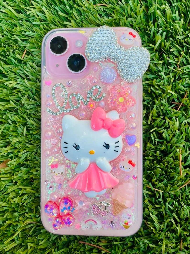 May include: Pink phone case with a Hello Kitty design. The case features a large, sparkling bow, the word "Love" in rhinestones, and various decorative elements like flowers, hearts, and an ice cream cone. The case is covered in glitter and has a clear border.
