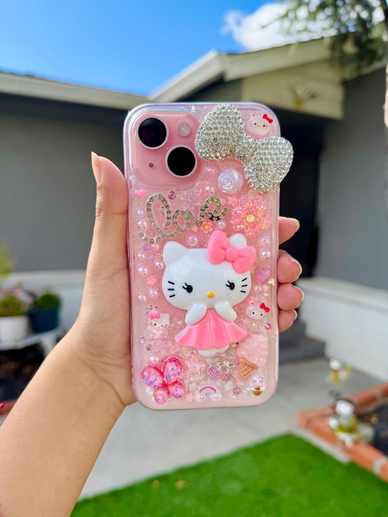 May include: Pink phone case featuring a Hello Kitty design with a large rhinestone bow, the word "Love", and various decorative elements. The case has a clear, glittery background and a pink border.
