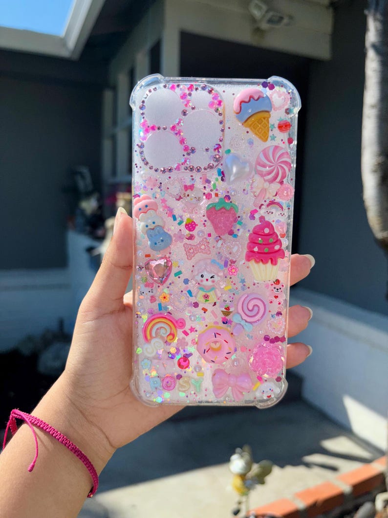 May include: Clear phone case filled with pink glitter and various charms, including an ice cream cone, a strawberry, a rainbow, and a donut. The case has a clear border and is held in a hand.