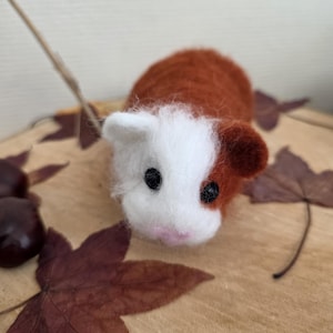 Peut inclure: Une figurine de cochon d'Inde en laine feutrée à l'aiguille, dotée d'une texture douce et duveteuse, avec un visage blanc et un pelage roux sur le corps. L'animal possède deux yeux en perles noires et un petit nez rose pâle en laine.