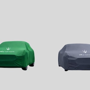 Puede incluir: Dos fundas para coche, una verde y otra gris, cada una cubriendo un coche. La funda verde tiene la palabra "Maserati" y un logotipo en blanco. La funda gris también tiene el logotipo y el texto "Maserati" en blanco. Ambos coches están sobre un fondo gris claro.