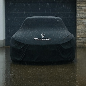 Puede incluir: Una funda negra para coche con el logotipo de Maserati en blanco, protegiendo un vehículo de la lluvia. La funda está colocada sobre el coche, con el emblema de Maserati centrado. El fondo incluye una puerta de garaje y una pared de ladrillo.
