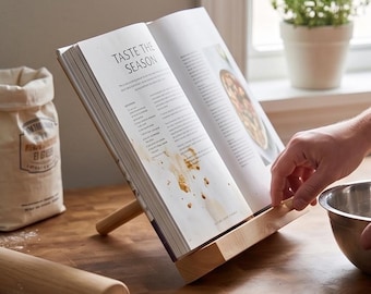 Op de afbeelding: Een kookboekstandaard van lichtgekleurd hout met een open kookboek waarop de tekst TASTE THE SEASON staat. De standaard heeft een schuine voet en een steunstang en toont een receptpagina met zichtbare vlekken en gedrukte tekst.