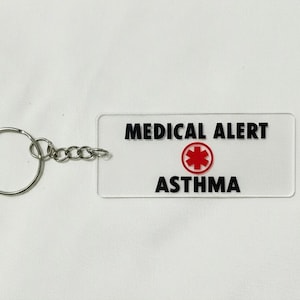 May include: A rectangular, clear acrylic keychain with the words "MEDICAL ALERT" and "ASTHMA" in black. A red medical symbol is centered between the words. The keychain has a silver metal ring and chain.