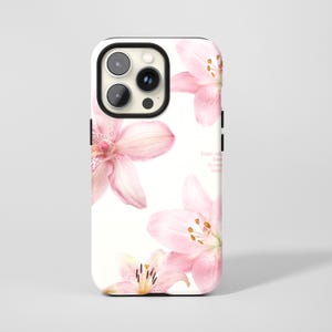 May include: White phone case featuring a floral design of pink lilies. The case has a black camera cutout and a quote in small, light pink text. The lilies have yellow stamens.