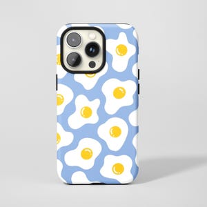 Könnte beinhalten: Eine Handyhülle mit hellblauem Hintergrund und einem sich wiederholenden Muster aus Spiegeleiern. Jedes Ei hat einen weißen Umriss und ein gelbes Eigelb. Die Hülle hat einen schwarzen Kamera- und Tastenbereich.