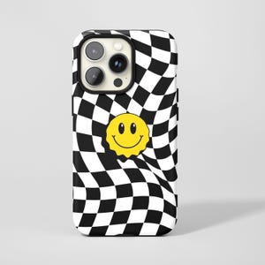 May include: A phone case with a black and white chequered design and a distorted appearance. A yellow smiley face is in the centre. The case has a black camera lens cutout.