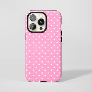 Puede incluir: Una funda de teléfono rosa con un patrón de lunares blancos. La funda tiene un borde negro alrededor de la cámara y botones laterales negros. La funda está diseñada para un teléfono inteligente.