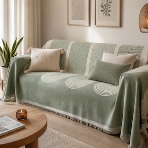 May include: A sage green sofa throw blanket featuring a repeating white arched line pattern and a fringed edge. The rectangular textile is draped over a sofa and is accompanied by a square cream-colored pillow and a rectangular sage green pillow.