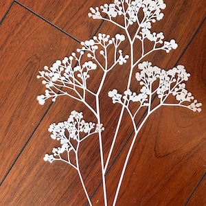 May include: White paper cut-out of a delicate flower sprig with multiple branches and small white blossoms. The sprig is on a brown wooden surface.