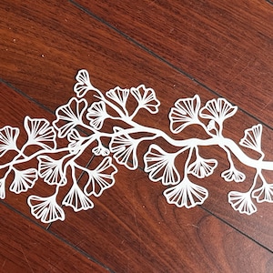 May include: White paper cut-out of a ginkgo tree branch with leaves. The branch is curved and has many leaves, all of which are detailed and intricate.